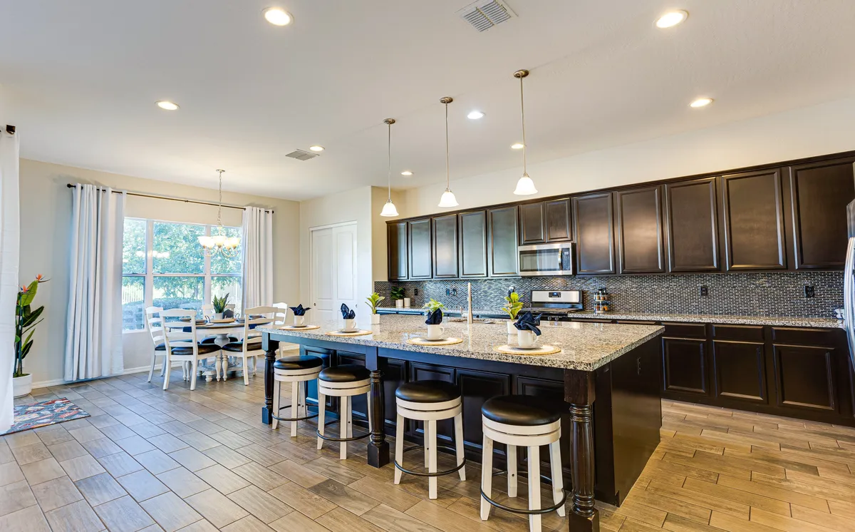 This image features a spacious kitchen with an island countertop, dark cabinetry, and a visible dining area. Large windows let in plenty of natural light, and the warm color palette creates an inviting atmosphere. The modern setup and open layout highlight both the kitchen’s functionality and its connection to the dining and social spaces.