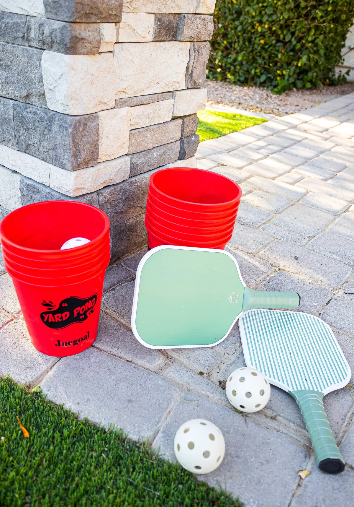 A lively yard pong game setup with large red buckets, green paddles, and white balls, positioned on a lush green lawn. The scene is energetic and playful, perfect for outdoor fun and friendly competition among guests.