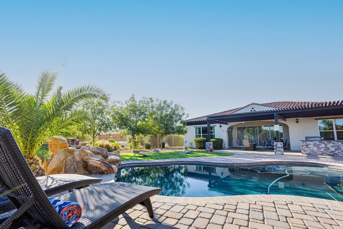 A tranquil backyard scene featuring a sparkling swimming pool, lounge chair, rock features, and palm trees, with the house exterior visible in the background. The area invites relaxation and offers ample space for unwinding after a busy day of activities.