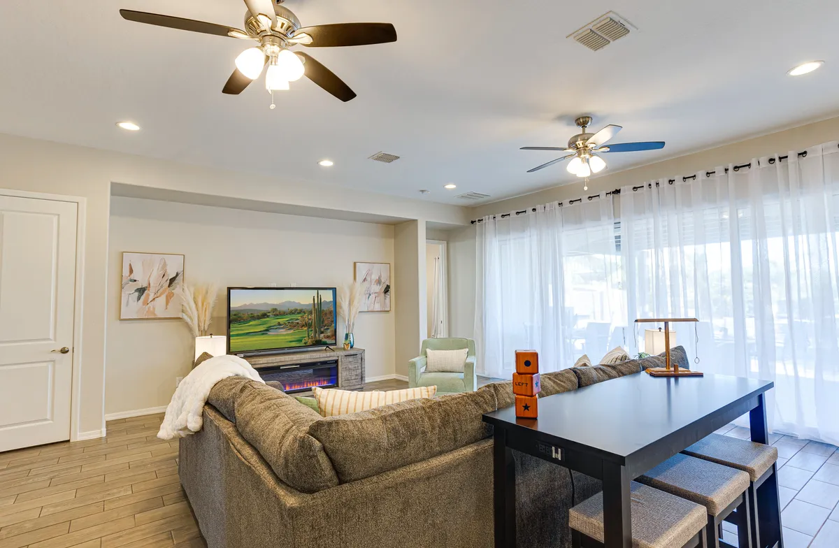 This image offers a sweeping view of a spacious open-concept living and dining area. It features a plush sectional sofa, a wall-mounted TV, a dining table with chairs, and expansive windows with sheer curtains letting in abundant natural light. Decorative artwork adds charm to the space, highlighting both the relaxation area and communal dining zone in one frame.