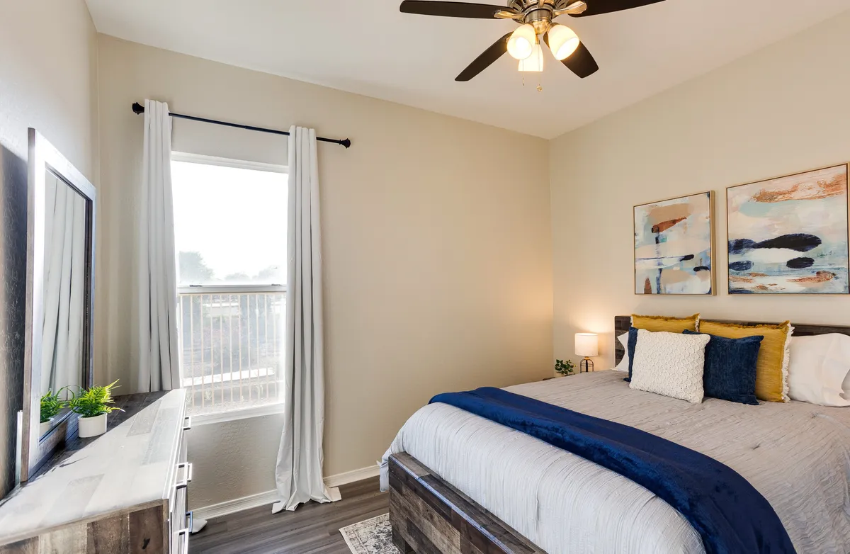 A cozy bedroom with a large window streaming in daylight, a bed dressed in blue and yellow accents, and decorative artwork that adds personality to the space. The overall ambiance is tranquil and inviting, suggesting a perfect haven for restful sleep.