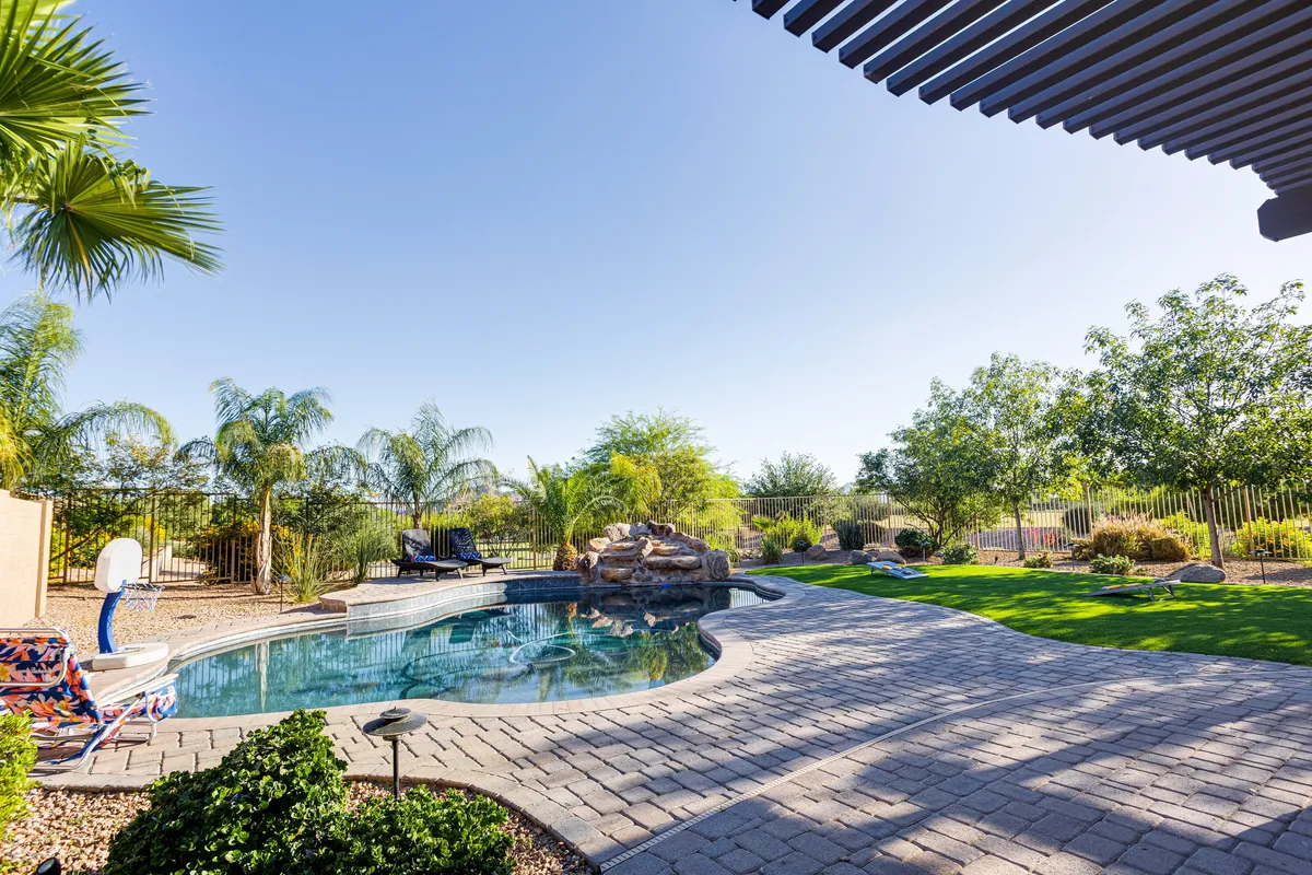 A tranquil pool scene featuring a rock formation, multiple lounge chairs, a landscaped garden, and a basketball hoop in the background. The setup is perfect for sunbathing, swimming, and socializing, with space for both relaxation and active fun.