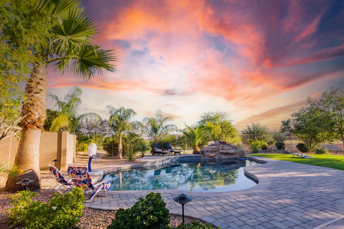 A serene backyard scene with a sparkling swimming pool bordered by colorful lounge chairs, lush greenery, and natural rock features. The inviting atmosphere suggests both relaxation and fun, with ample space for sunbathing and poolside gatherings.